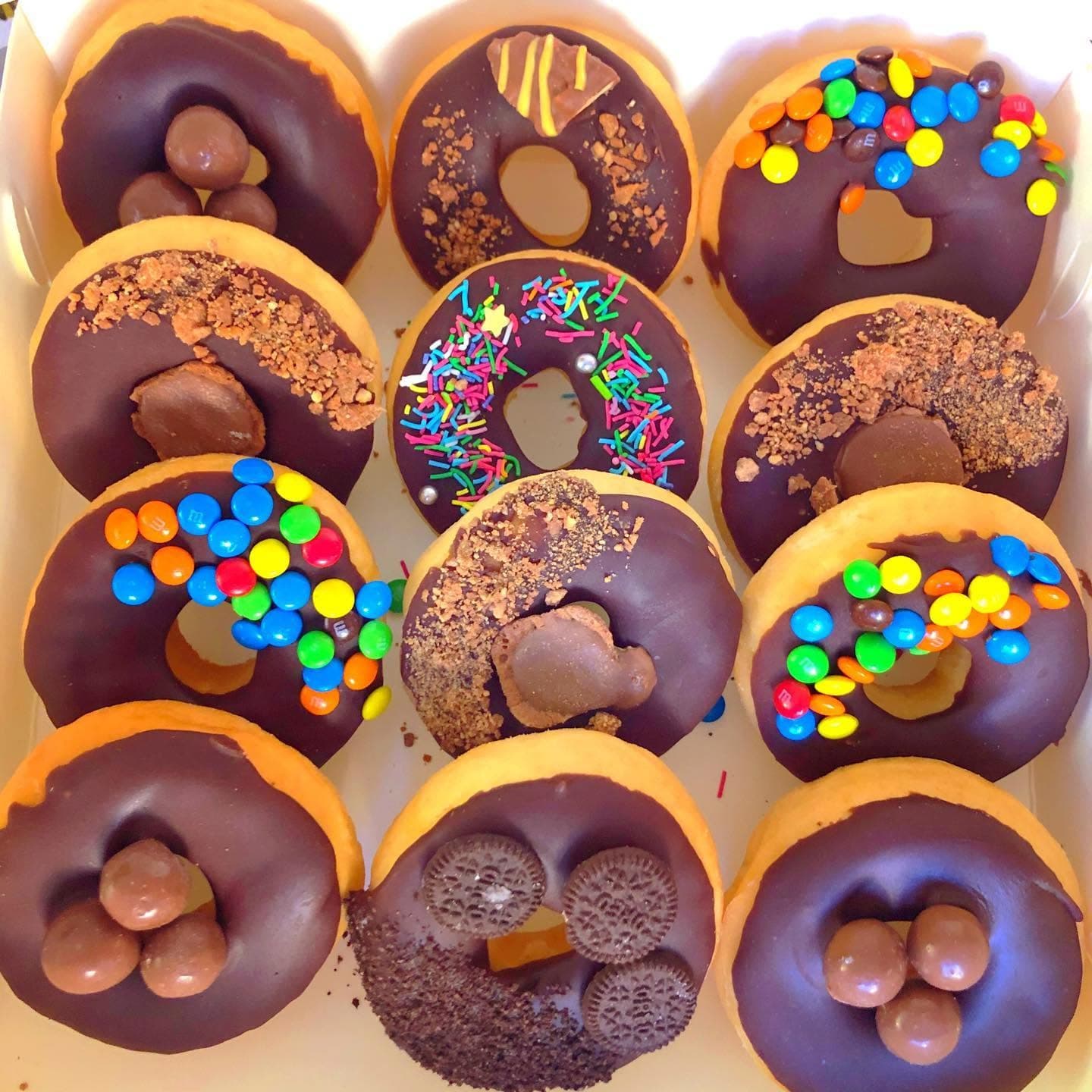 A box of chocolate-frosted donuts topped with M&Ms, sprinkles, Maltesers, and crushed cookies.
