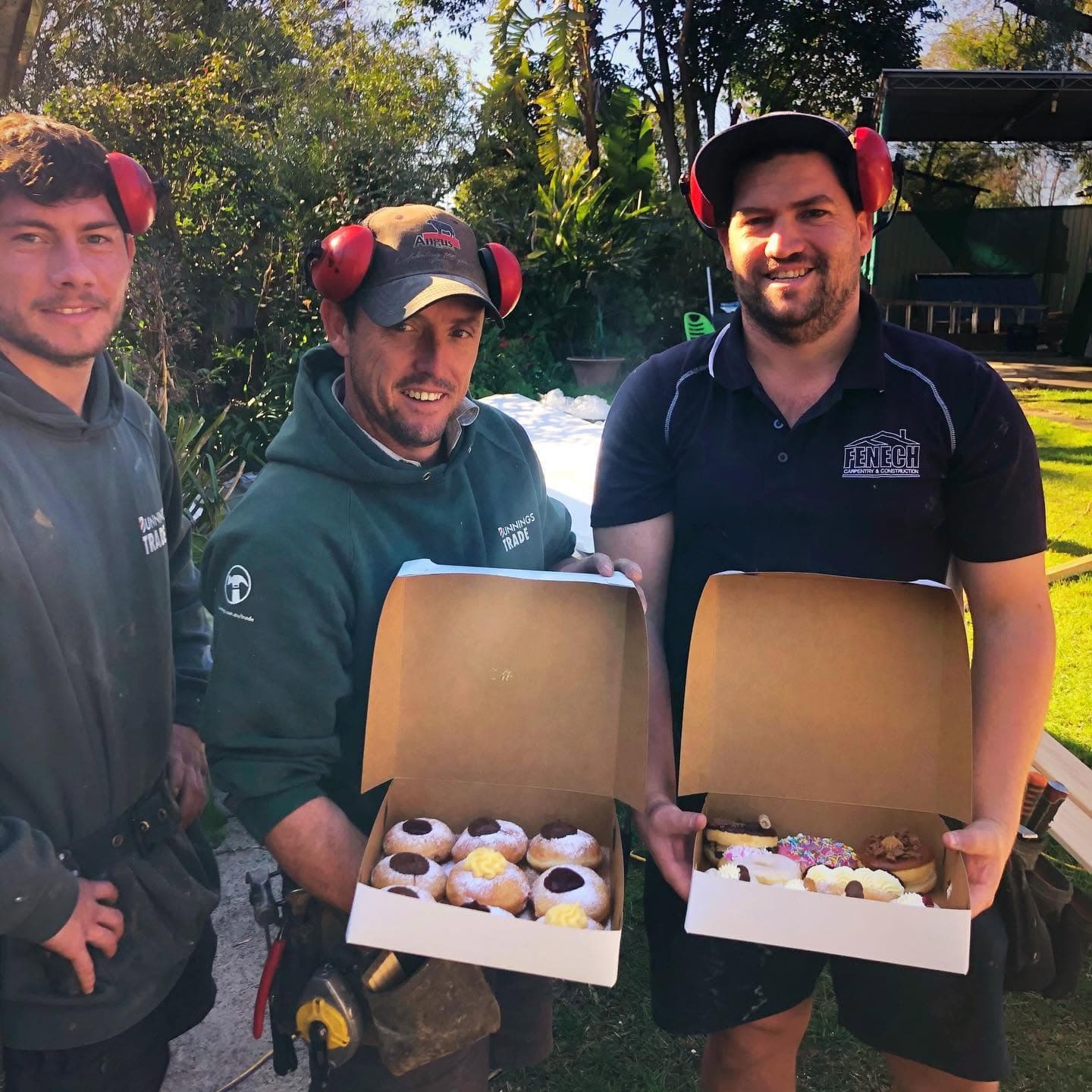 Three construction workers wearing red earmuffs smile and hold open boxes of assorted donuts.