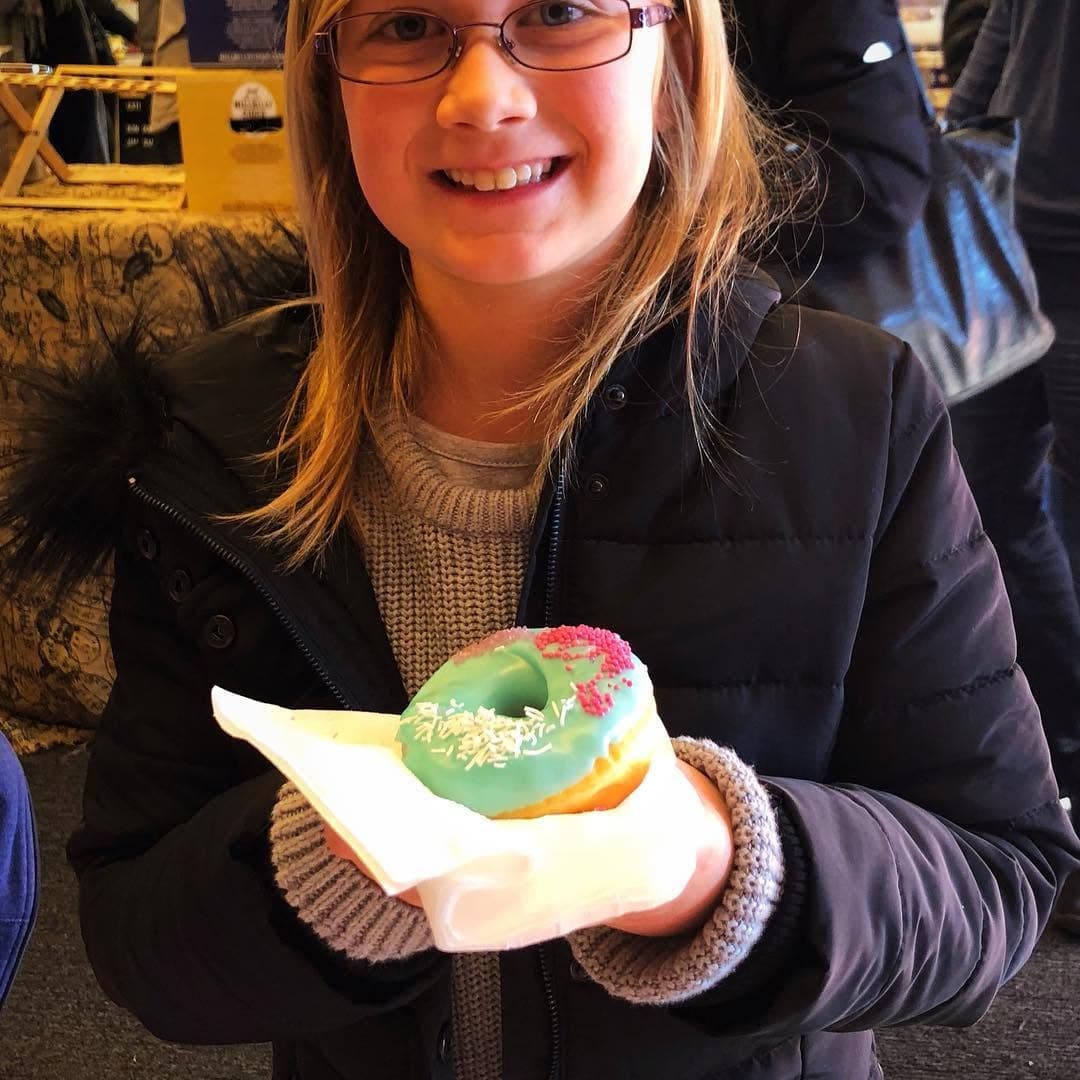 Smiling young girl with glasses holding a light blue frosted donut with colorful sprinkles.