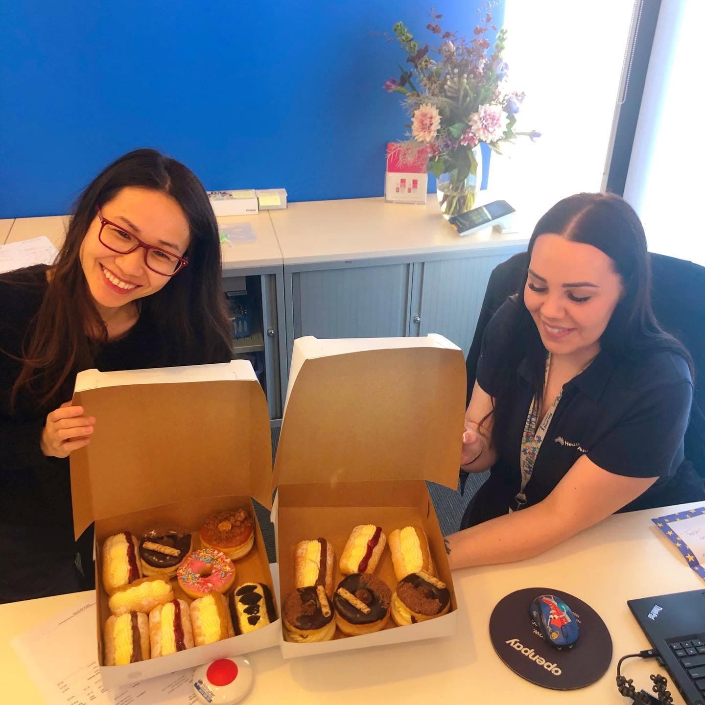 Two smiling women in an office display two open boxes of assorted colorful donuts.