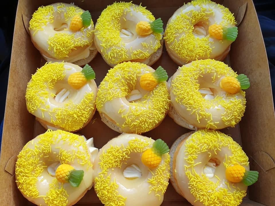 Nine glazed donuts with yellow sprinkles and gummy pineapple candies in a cardboard bakery box.