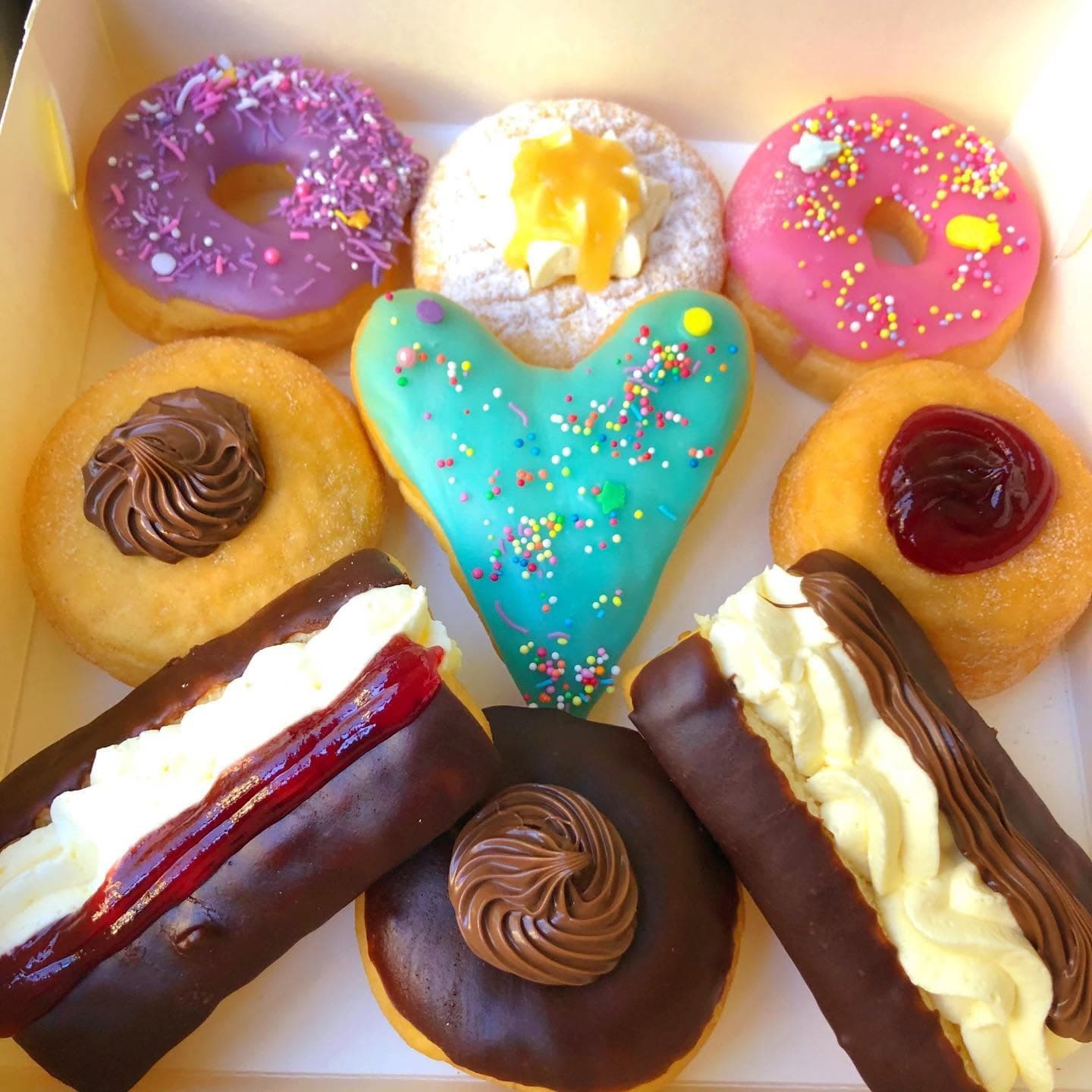 Assorted colorful donuts and cream-filled pastries, featuring a teal heart-shaped donut in a box.