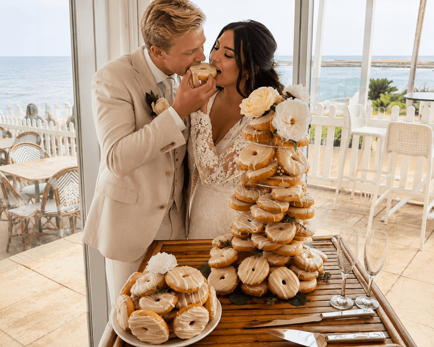 Newlyweds sharing a donut beside a tiered donut tower at their bright seaside wedding reception.